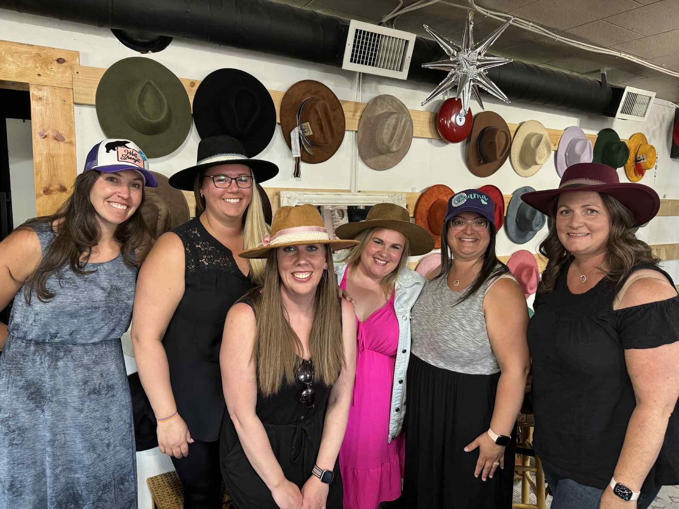Six guests posing in custom styled hats in front of the hat wall