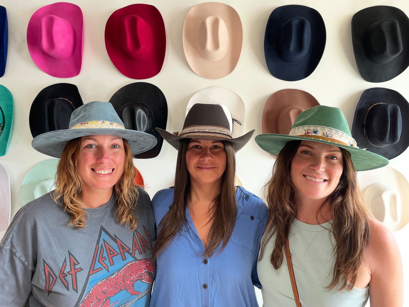Three guests posing in custom styled hats in front of the hat wall