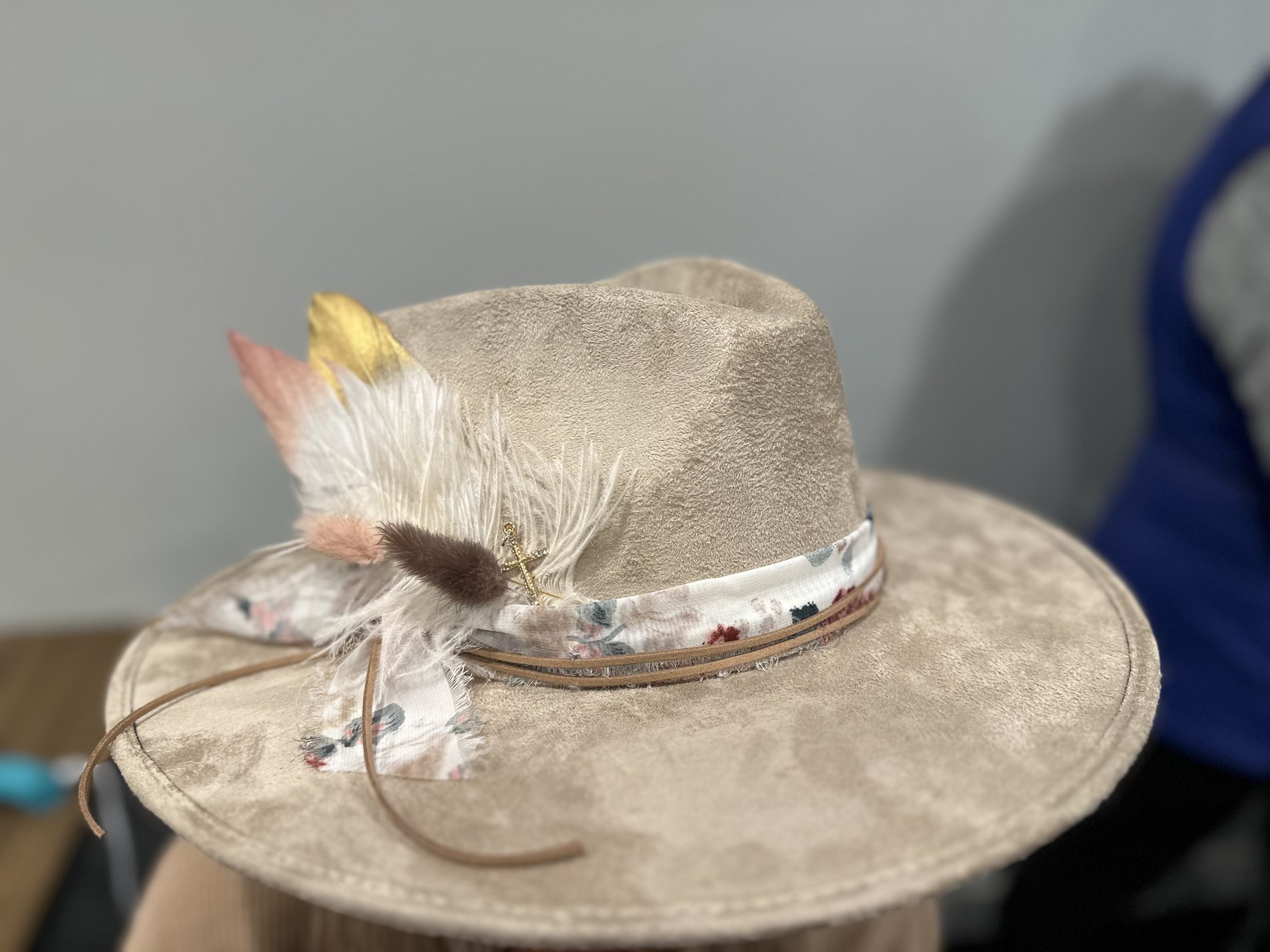 Close-up of a custom cream hat styled with feathers, a floral band, and a gold cross charm at a Royal LePage event