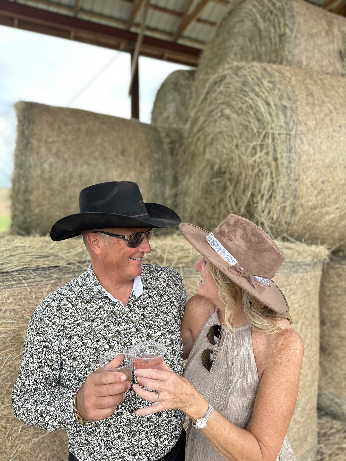 A couple toasting in their custom cowboy hats inside a barn, hay bales behind them
